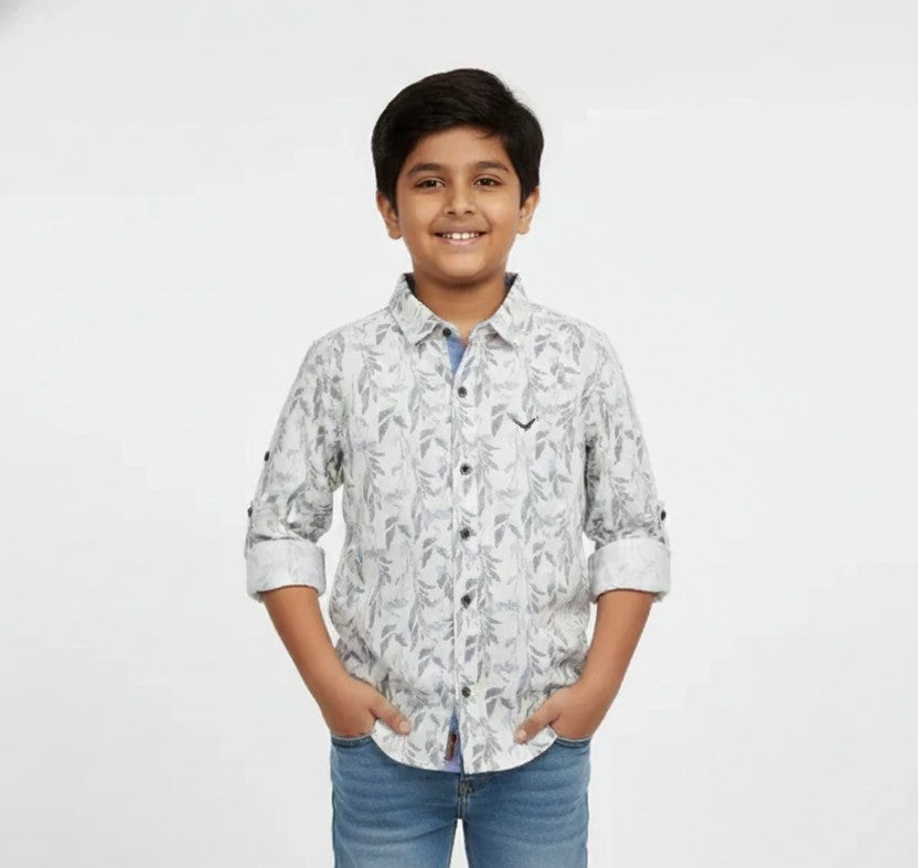 A young boy smiling with his hands in his pockets, modeling a Fyst brand light grey button-down shirt featuring an all-over delicate leaf and branch print. The shirt includes contrasting dark buttons, a small chest logo, and sleeves with roll-up tab details, set against a solid white background.