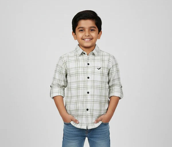 A young boy smiling with his hands in his pockets, modeling a Fyst brand off-white button-down shirt featuring a green and grey windowpane check pattern. The shirt has dark contrasting buttons, a small black logo on the chest, and long sleeves with roll-up tab details, shown against a neutral studio background.