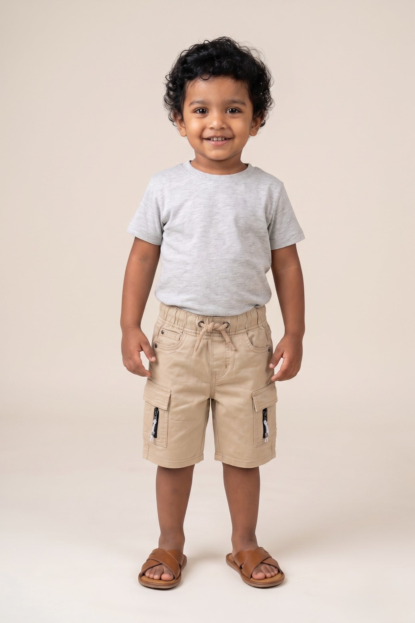 A kid wearing FYST brand lightweight khaki cargo shorts featuring an elasticated drawstring waistband, side flap pockets with decorative zipper details, and a comfortable knee-length fit.