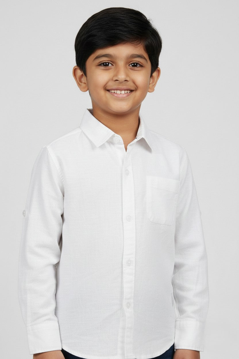 A smiling young boy wearing a Fyst brand solid white long-sleeve cotton shirt. The shirt features a classic spread collar, matching white buttons, a single chest pocket, and a clean, textured fabric finish. He is posed against a light grey background, showcasing the shirt's bright, formal look and high-quality tailoring.