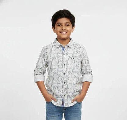 A young boy smiling with his hands in his pockets, modeling a Fyst brand light grey button-down shirt featuring an all-over delicate leaf and branch print. The shirt includes contrasting dark buttons, a small chest logo, and sleeves with roll-up tab details, set against a solid white background.