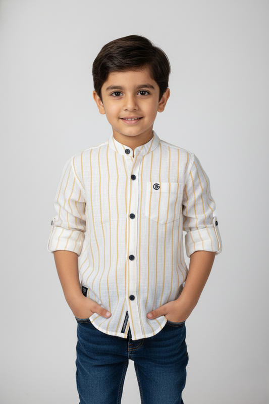 A front-facing studio image of a young boy wearing a Fyst brand off-white button-down shirt with vertical yellow pinstripes. The shirt features a mandarin collar, contrasting dark buttons, a single chest pocket, and rolled sleeves secured with button tabs. He is wearing blue jeans and posing with hands in pockets against a light grey background.