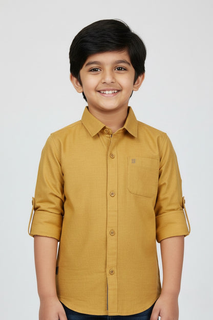 A smiling young boy wearing a Fyst brand mustard yellow long-sleeve cotton shirt. The shirt features a classic spread collar, color-matched buttons, a single chest pocket, and sleeves rolled up with button tabs. He is posed against a plain white background, highlighting the shirt's bold color and textured premium fabric.