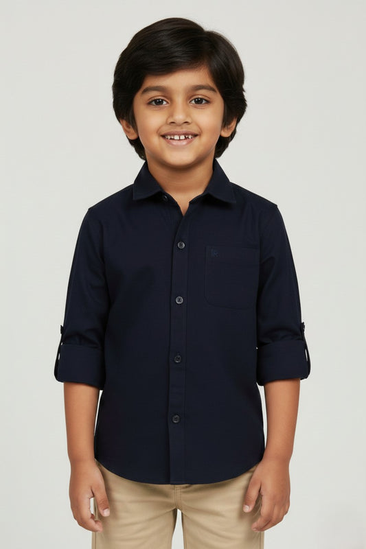 A smiling young boy wearing a Fyst brand solid navy blue long-sleeve cotton shirt. The shirt features a classic spread collar, matching navy buttons, a single chest pocket, and sleeves rolled up with button tabs. He is paired with khaki trousers and posed against a light grey background, showcasing the shirt's sharp tailoring and deep, professional blue color.