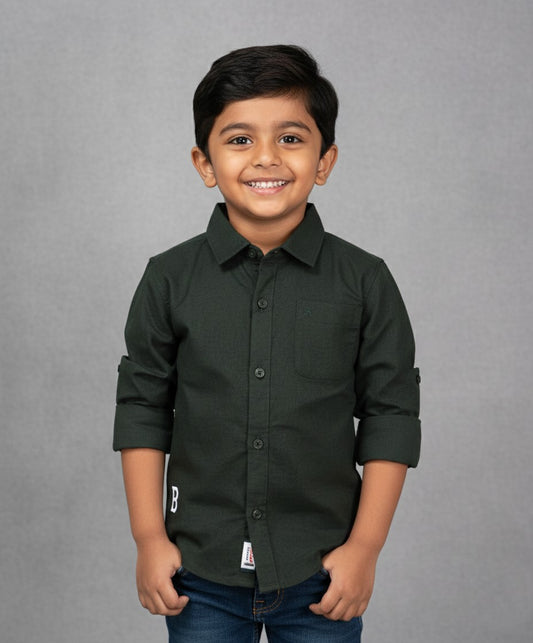 A smiling young boy models a Fyst brand solid olive green long-sleeve cotton shirt. The shirt features a classic spread collar, color-matched dark buttons, a single chest pocket, and sleeves rolled up with button tabs. He is posed against a textured grey background, showcasing the shirt's rich earth-tone color and durable fabric.
