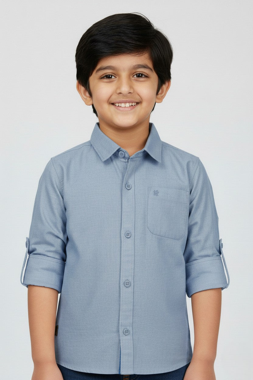 A smiling young boy wearing a Fyst brand slate blue long-sleeve cotton shirt. The shirt features a classic spread collar, matching blue buttons, a single chest pocket, and sleeves rolled up with button tabs. He is posed against a plain white background, highlighting the shirt's smooth fabric texture and modern, dusty blue color.