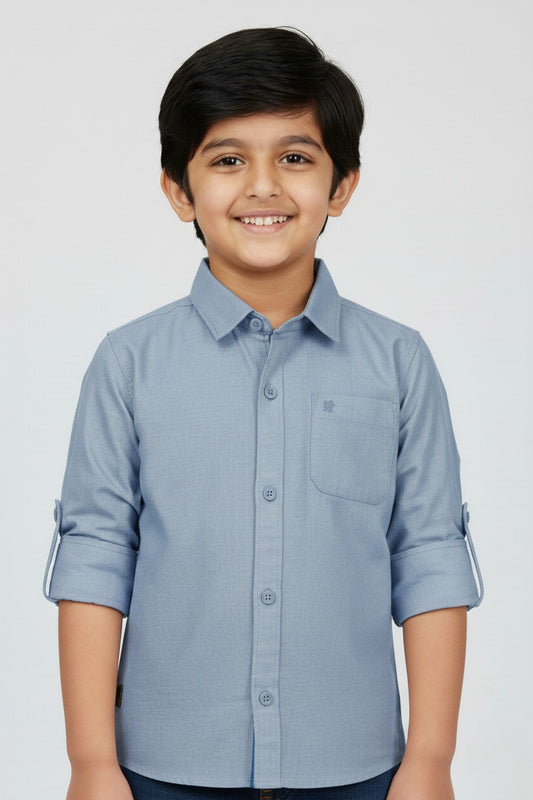 A smiling young boy wearing a Fyst brand slate blue long-sleeve cotton shirt. The shirt features a classic spread collar, matching blue buttons, a single chest pocket, and sleeves rolled up with button tabs. He is posed against a plain white background, highlighting the shirt's smooth fabric texture and modern, dusty blue color.