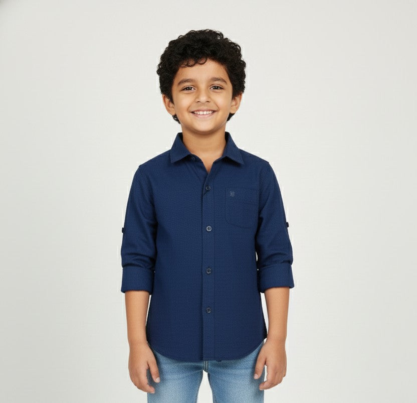 A young boy with curly hair smiling in a studio setting, wearing a Fyst brand deep navy blue button-down shirt. The shirt features a subtle textured fabric, a single chest pocket, and sleeves rolled up with button-tab details, presented against a plain light grey background.