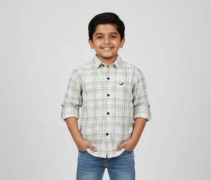 A young boy smiling with his hands in his pockets, modeling a Fyst brand off-white button-down shirt featuring a green and grey windowpane check pattern. The shirt has dark contrasting buttons, a small black logo on the chest, and long sleeves with roll-up tab details, shown against a neutral studio background.