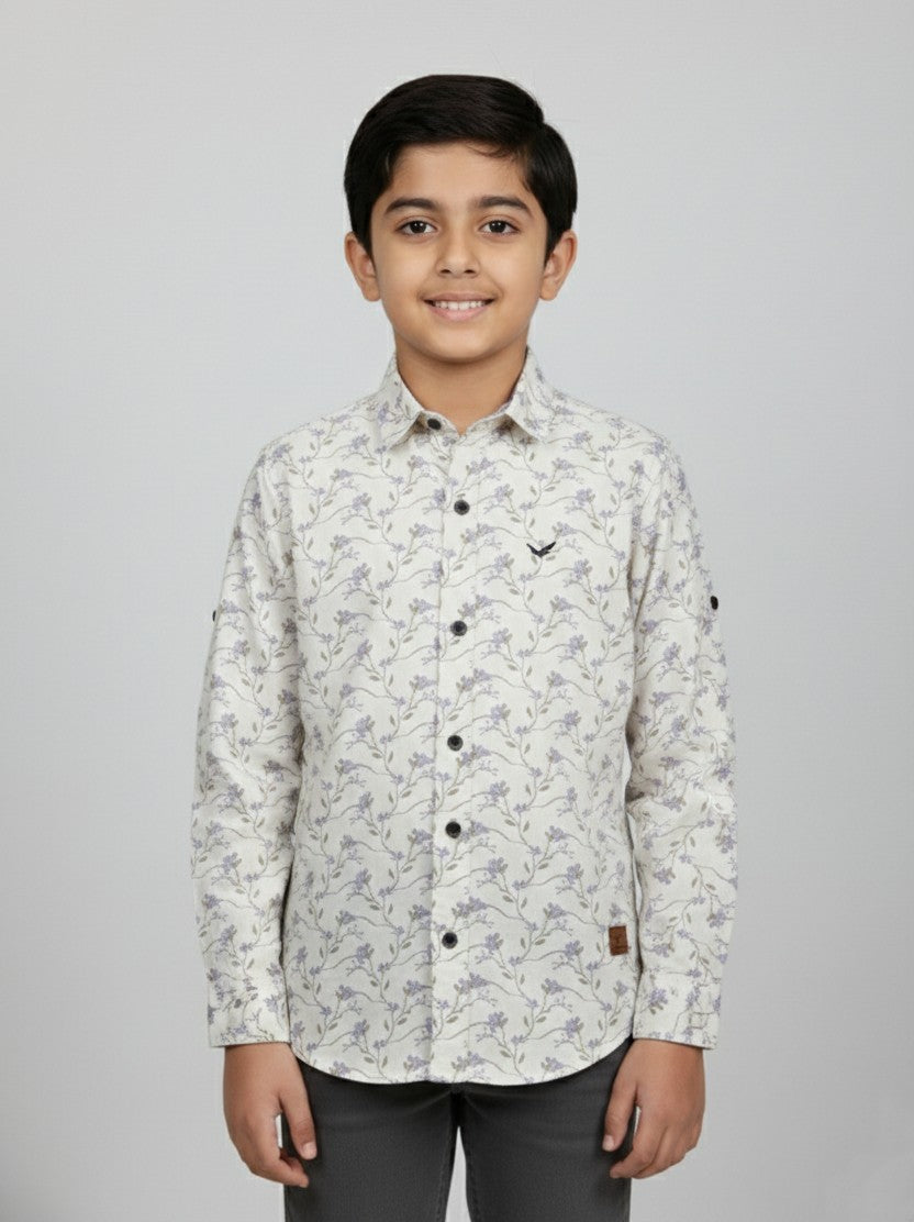 A young boy smiling in a studio portrait, modeling a Fyst brand cream-colored button-down shirt featuring a delicate all-over lavender floral and vine print. The shirt includes contrasting dark buttons, a small chest logo, and versatile roll-up sleeves with button tabs, shown against a solid light grey background.
