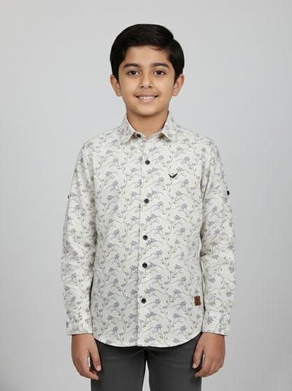 A young boy smiling in a studio portrait, modeling a Fyst brand cream-colored button-down shirt featuring a delicate all-over lavender floral and vine print. The shirt includes contrasting dark buttons, a small chest logo, and versatile roll-up sleeves with button tabs, shown against a solid light grey background.