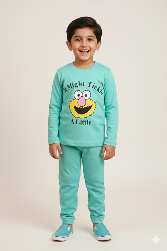 A young boy modeling a matching aqua blue long-sleeve pajama set. The top features a whimsical yellow character face with the playful text "It Might Tickle A Little." He is standing against a neutral background wearing matching aqua blue slip-on shoes.