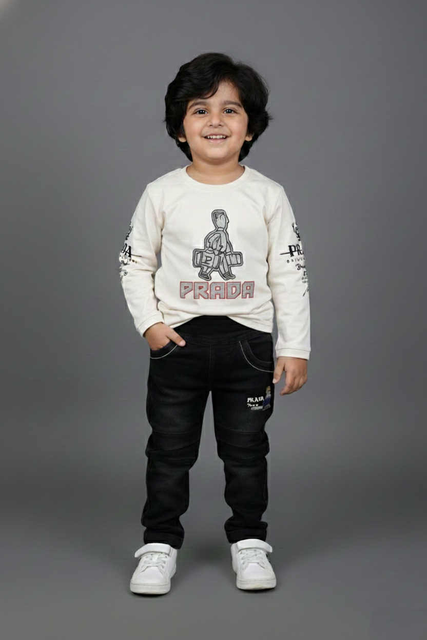 A young boy modeling a stylish two-piece outfit. He is wearing an off-white long-sleeve t-shirt featuring a central character graphic and the word "PRIDE" in red, paired with slim-fit black washed denim jeans. He is posing against a neutral grey background with white sneakers.