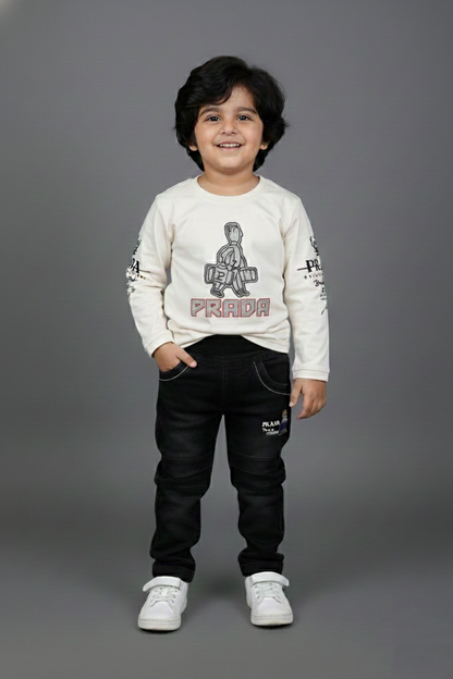 A young boy modeling a stylish two-piece outfit. He is wearing an off-white long-sleeve t-shirt featuring a central character graphic and the word "PRIDE" in red, paired with slim-fit black washed denim jeans. He is posing against a neutral grey background with white sneakers.