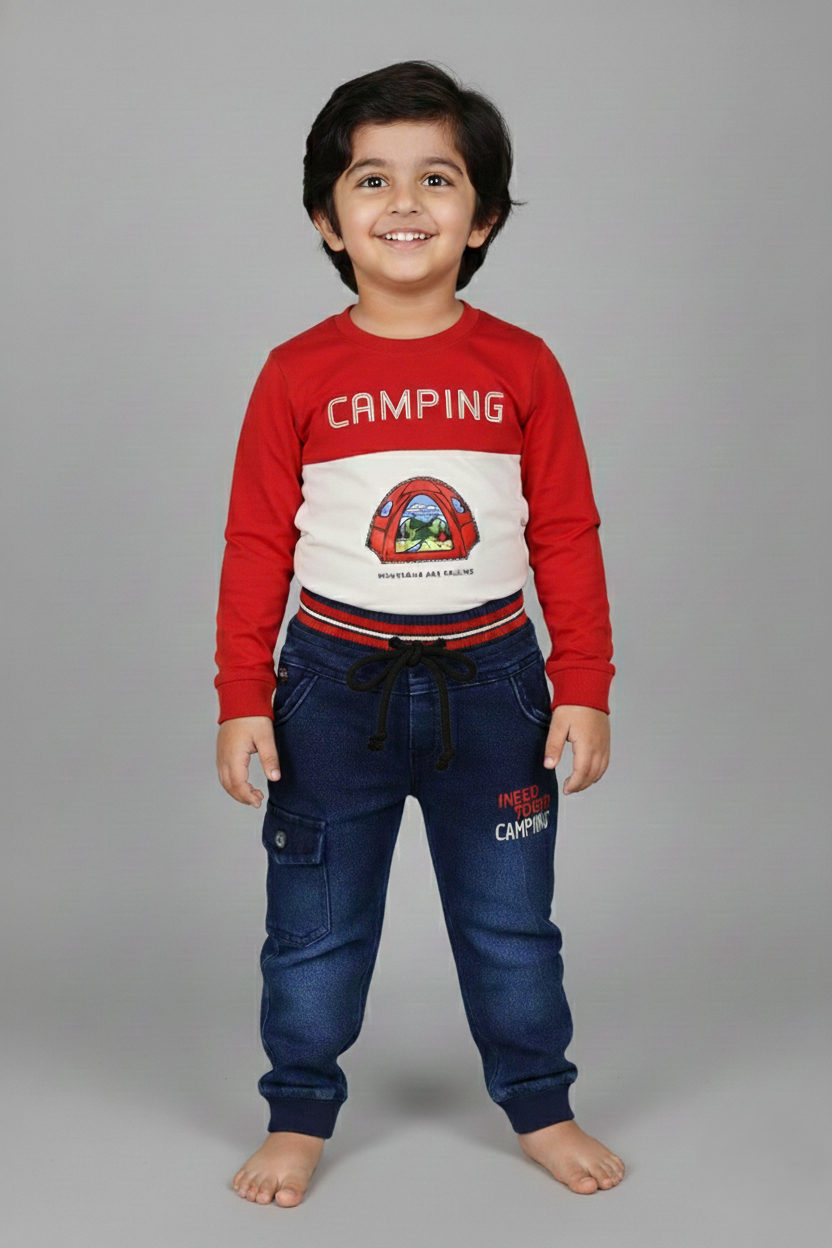 A young boy modeling a two-piece outdoor-themed set. The outfit features a long-sleeve t-shirt with a red top half and cream bottom half, printed with a colorful "Camping" mountain graphic. He is wearing matching dark blue denim joggers that have a red and white striped elastic waistband and a small cargo pocket on the leg.