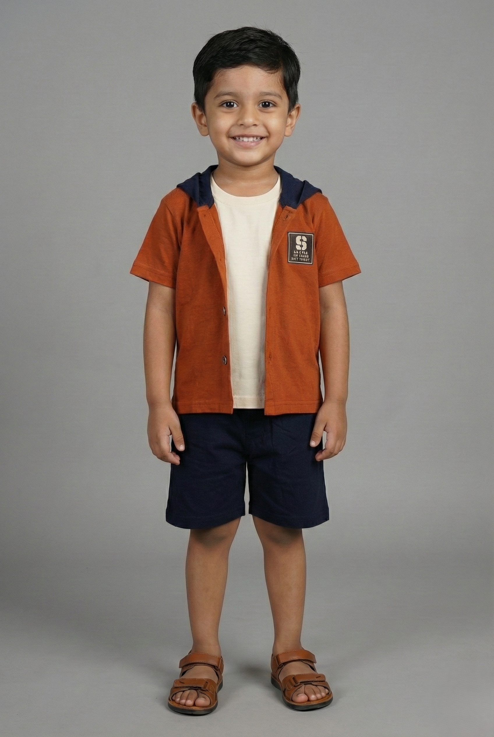 A young boy modeling a trendy three-piece summer outfit. The set includes a rust orange short-sleeve button-down shirt with a navy blue hood, worn over a plain cream t-shirt. He is wearing matching navy blue shorts and brown leather sandals, standing against a neutral studio background.