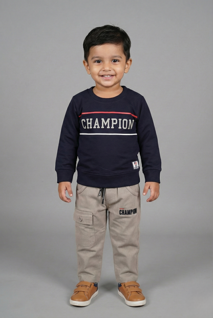 A young boy modeling a two-piece winter clothing set. The navy blue long-sleeve sweatshirt features a white "CHAMPION" graphic across the chest with a thin red accent stripe. He is wearing matching light beige cargo joggers with a functional side pocket and brown sneakers.