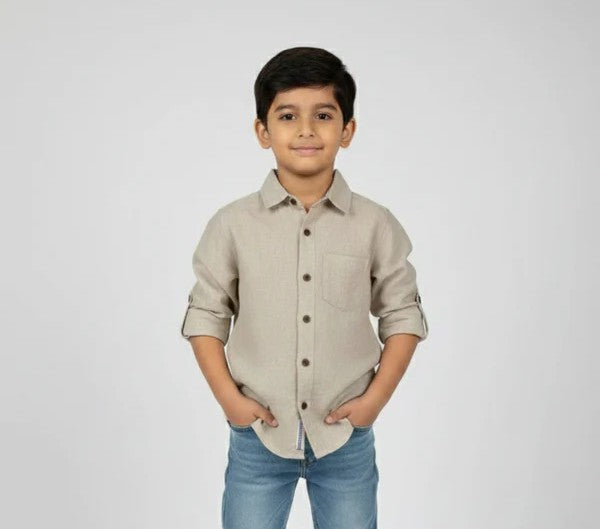 A young boy smiling with his hands in his pockets, modeling a Fyst brand beige linen-textured button-down shirt. The shirt features a classic collar, a single chest pocket, dark brown buttons, and long sleeves with roll-up tab details, shown against a neutral studio background.