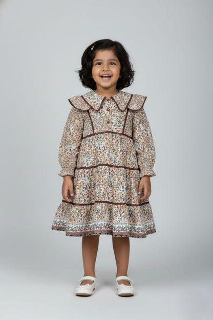 "A long-sleeved, tiered girls' dress featuring a dense floral print in autumn tones of brown, teal, and rust on a cream background. The dress has a bohemian style with ruffle details on the shoulders and dark maroon lace trim accentuating the yoke, waist, and skirt tiers. It features a button-down front placket with reddish-brown buttons and elasticized cuffs on the sleeves