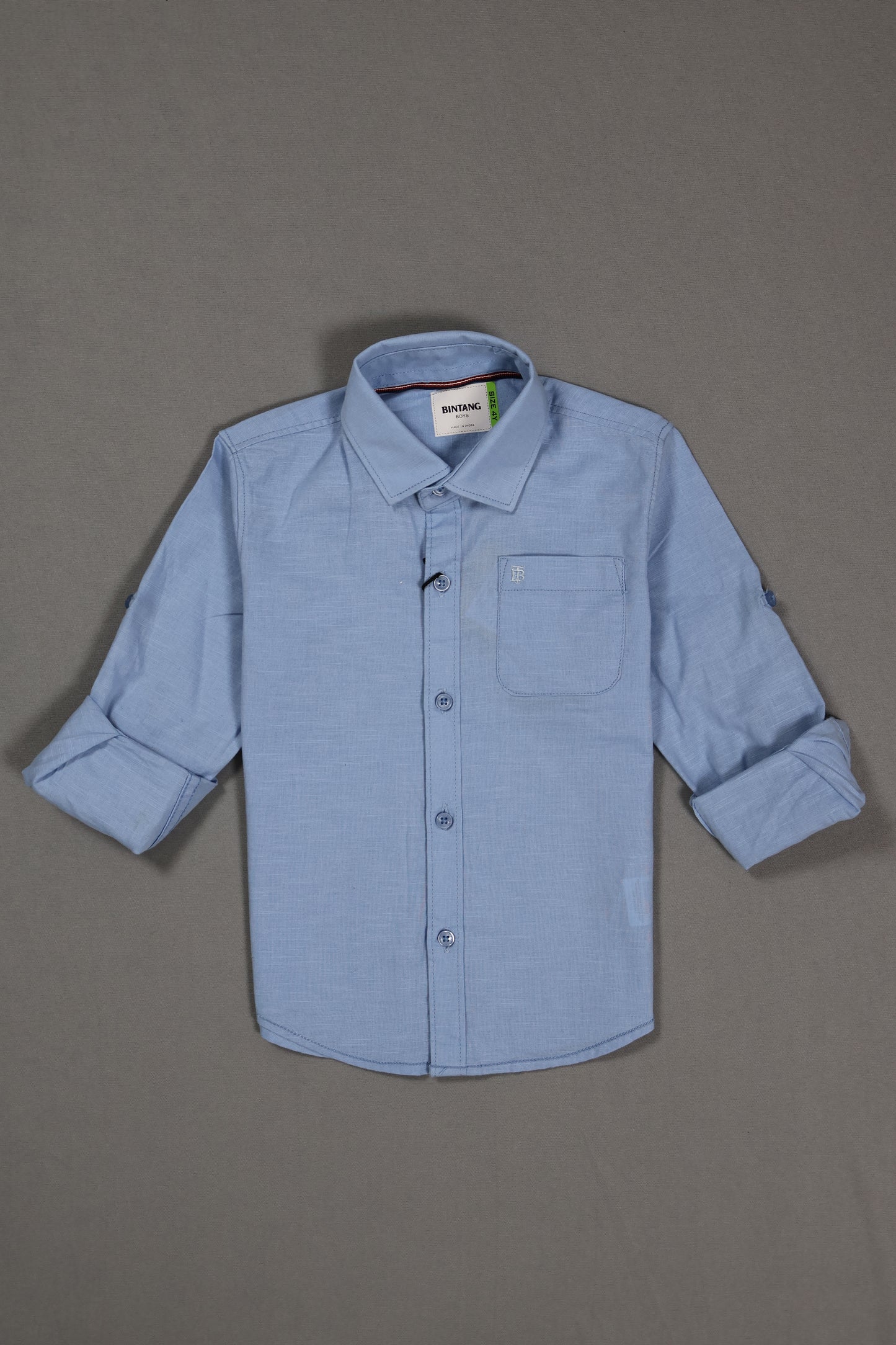 Light blue cotton kids shirt with long sleeves and front pocket, laid flat on gray background
