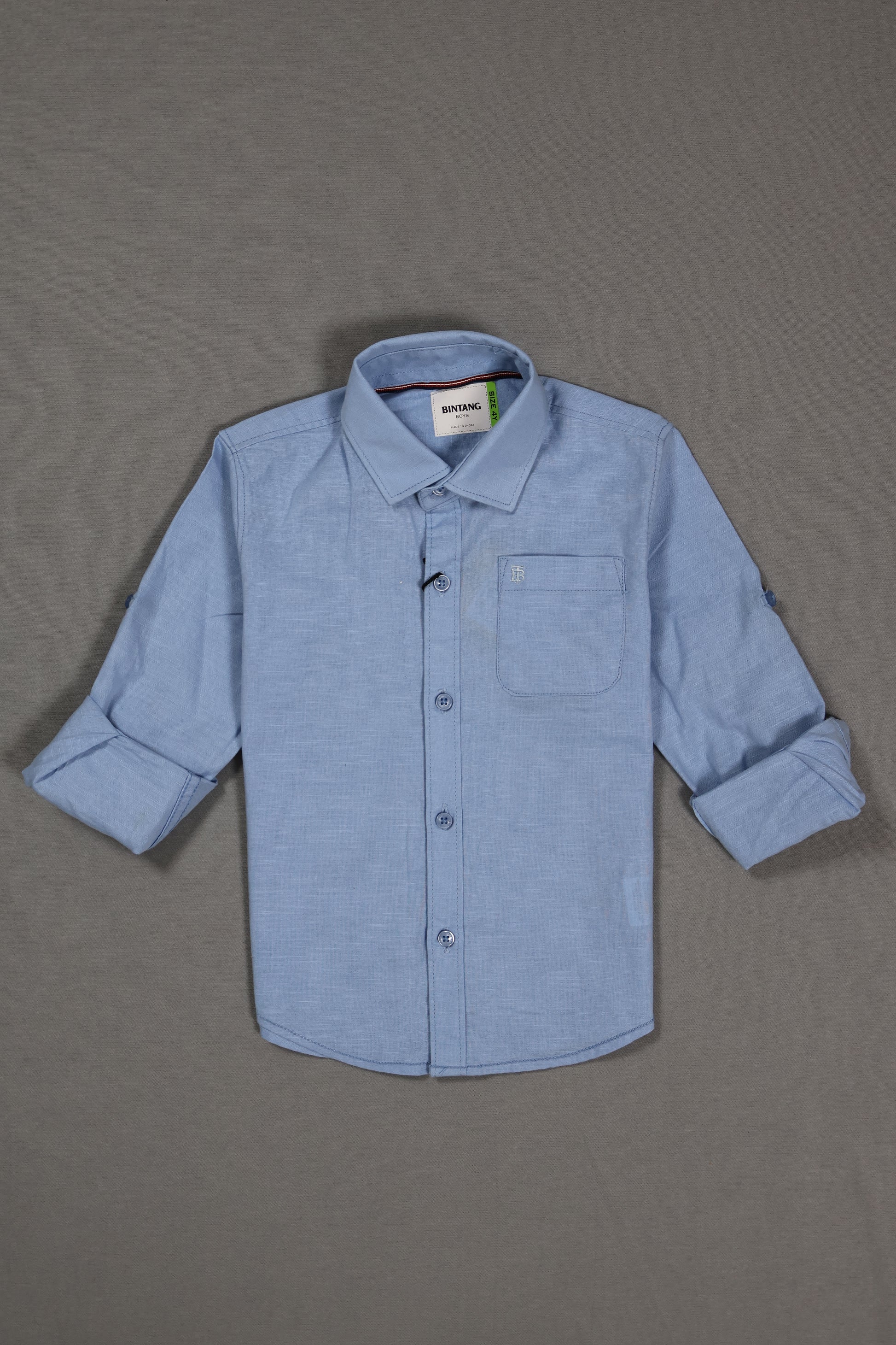 Light blue cotton kids shirt with long sleeves and front pocket, laid flat on gray background