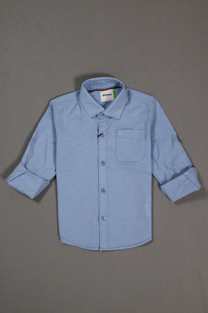 Light blue cotton kids shirt with long sleeves and front pocket, laid flat on gray background