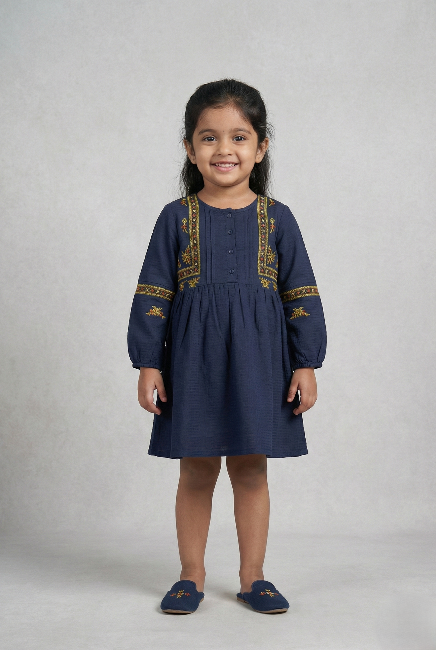 "A navy blue, long-sleeved girls' dress with a button-down bodice. It features vertical bands of yellow, orange, and red geometric and floral embroidery framing the buttons, with matching horizontal embroidered bands across the sleeves. The fabric has a subtle textured weave, and the skirt is gathered at the waist