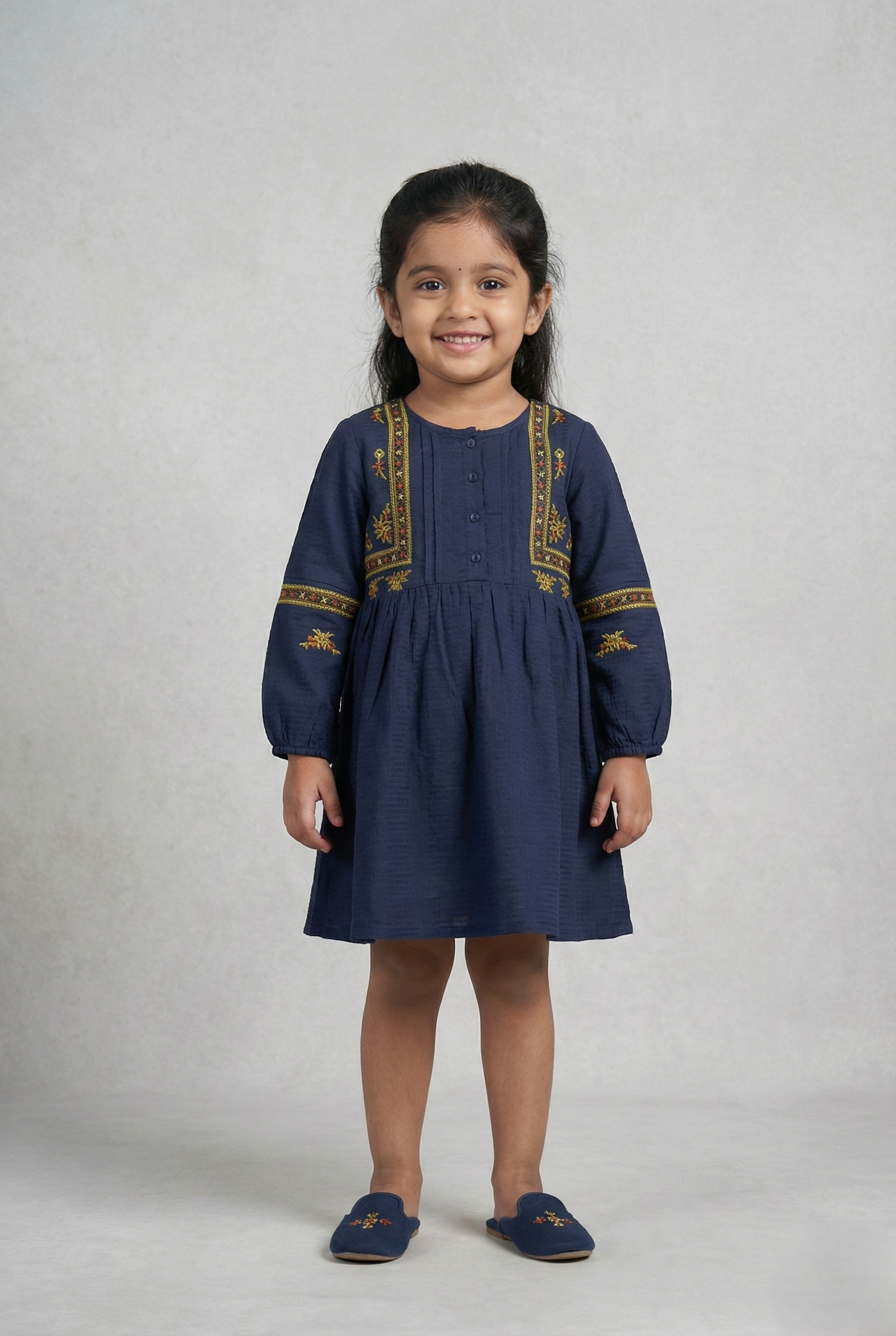 "A navy blue, long-sleeved girls' dress with a button-down bodice. It features vertical bands of yellow, orange, and red geometric and floral embroidery framing the buttons, with matching horizontal embroidered bands across the sleeves. The fabric has a subtle textured weave, and the skirt is gathered at the waist