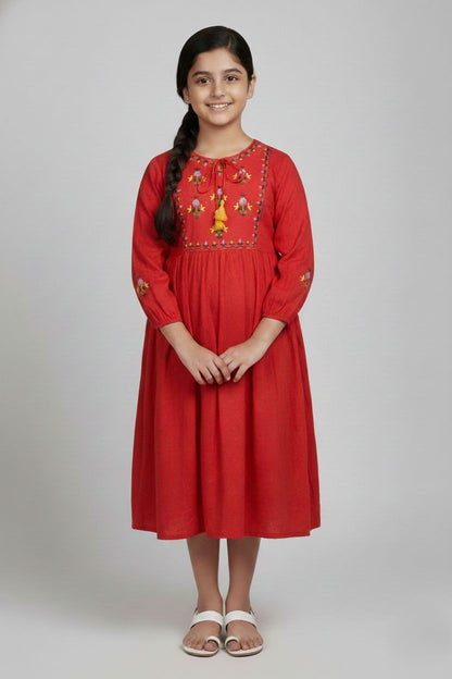 A bright red, long-sleeved girls' dress hanging on a wooden hanger. It features a square bodice area with yellow and light blue floral embroidery and dark stitching. The dress includes a round neckline with a drawstring tie ending in yellow tassels, gathered sleeve cuffs with matching embroidery, and a flowy skirt