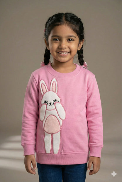 A pink long-sleeved sweatshirt for girls featuring a large, textured appliqué of a white bunny on the front. The bunny has pink inner ears and a pink belly, depicted with its paws raised to its cheeks in a cute pose. The sweatshirt has a ribbed crew neck, ribbed cuffs, and a ribbed hem
