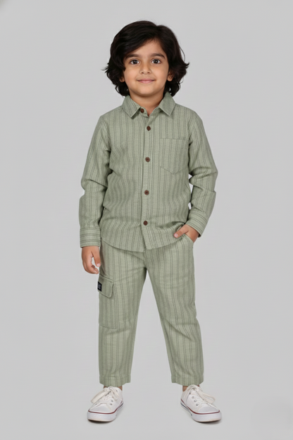 "A detailed product showcase featuring the Fyst boys' pistachio green striped co-ord set. The primary image shows a young boy modeling the complete set, which includes a long-sleeved button-down shirt with fine vertical pinstripes and matching straight-cut trousers. The shirt features a classic spread collar, a single chest pocket, and prominent dark red contrast buttons. The accompanying thumbnails highlight the individual pieces, specifically showing the trousers with a comfortable elasticated waistband a